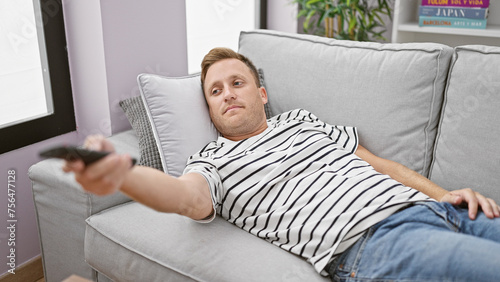 Bored young caucasian man, lounging on the sofa, hopelessly channel surfing at home