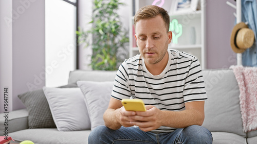 Handsome young caucasian man, focused and relaxed, using his smartphone while sitting on a sofa in living room, immersed in the world of technology and internet, at his cozy apartment interior.