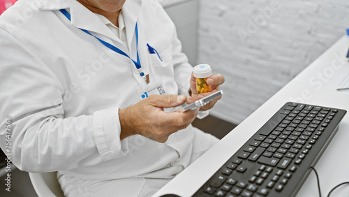 A mature man in a lab coat examines medication in a hospital setting with a smartphone and keyboard nearby. © Krakenimages.com