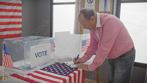 A mature man in a pink shirt writes at a polling station with us flags and a ballot box indicating a voting scene. photo