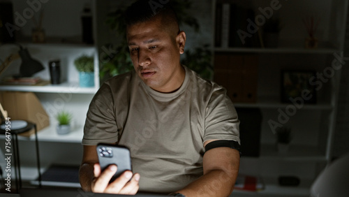 Photos Night time hustle, young, serious latin man, a hard-working business professional, immersed in texting on his smartphone at the office desk