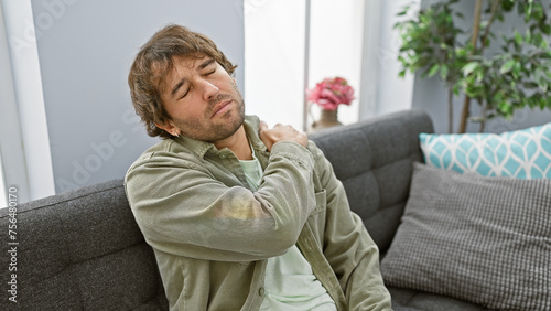 Young man with beard in pain holding neck while sitting on a couch indoors.