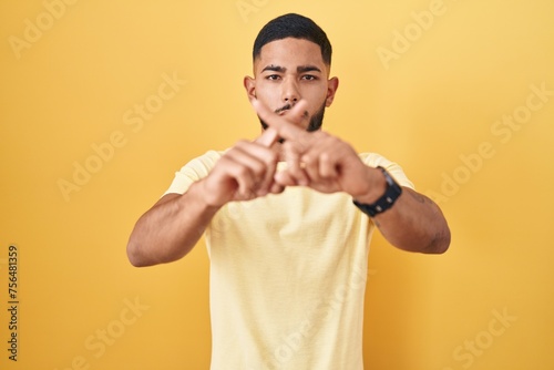 Young hispanic man standing over yellow background rejection expression crossing fingers doing negative sign