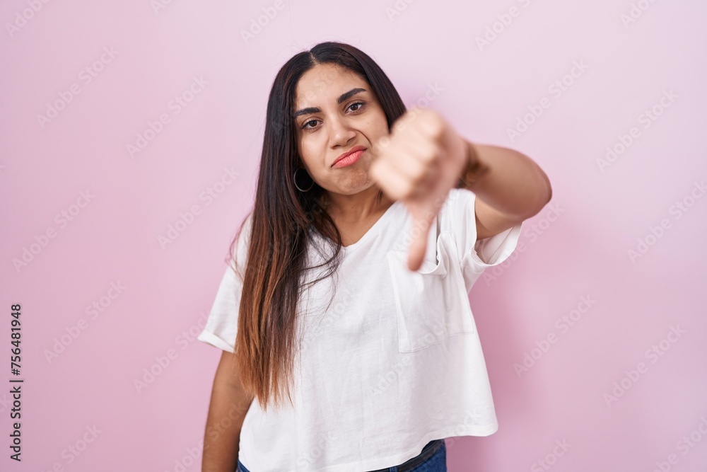 Young arab woman standing over pink background looking unhappy and angry showing rejection and negative with thumbs down gesture. bad expression. Young arab woman standing over pink background looking unhappy and angry showing rejection and negative with thumbs down gesture. bad expression.