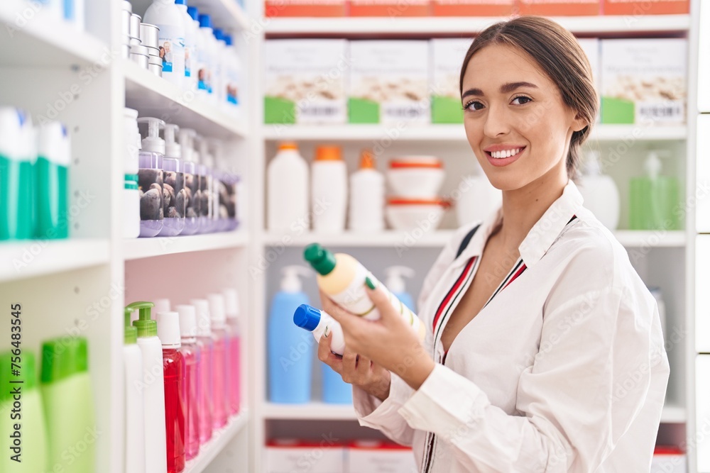 Young beautiful hispanic woman customer smiling confident holding shampoo bottles at pharmacy
