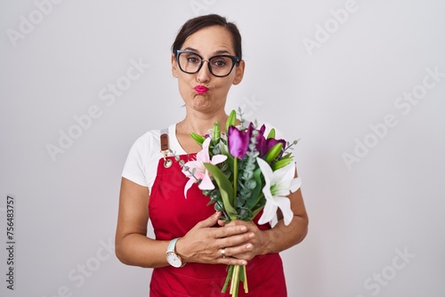 Middle age brunette woman wearing apron working at florist shop holding bouquet puffing cheeks with funny face. mouth inflated with air, crazy expression.