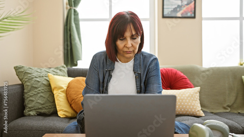 Focused senior hispanic woman using laptop in a cozy living room adorned with colorful cushions.