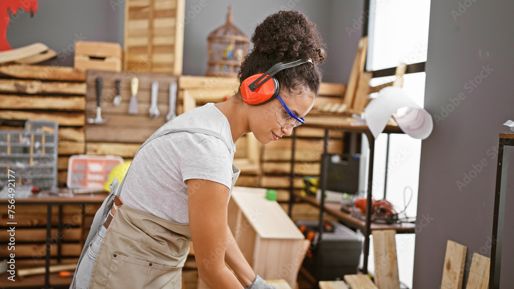 Attractive hispanic female carpenter smiling at her carpentry workshop, young, beautiful woman with curly hair, glasses and headphones bringing joy to the woodworking industry