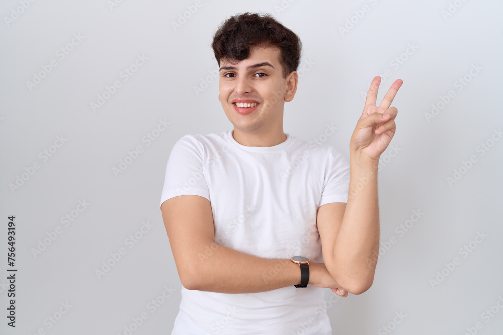 Young non binary man wearing casual white t shirt smiling with happy face winking at the camera doing victory sign with fingers. number two. Young non binary man wearing casual white t shirt smiling with happy face winking at the camera doing victory sign with fingers. number two.