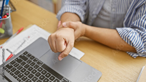 A man in an office winces in pain, clutching his wrist over a laptop, suggesting carpal tunnel syndrome or injury.