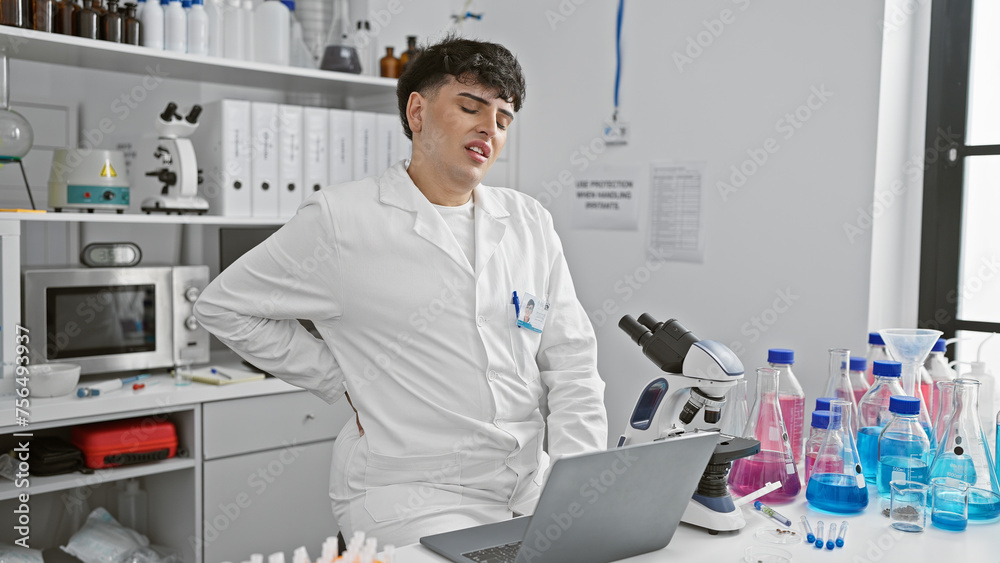 A scientist man in a laboratory suffering from back pain while working on a research project with a microscope and laptop.