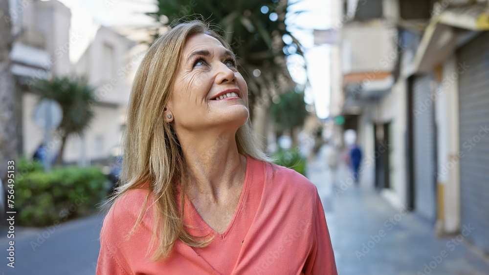 A smiling middle-aged blonde woman looks up as she strolls outdoors on a city street, exuding a sense of joy and contentment.
