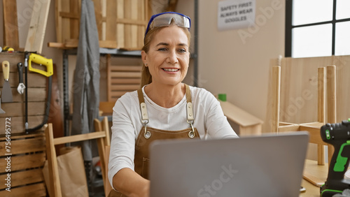 A smiling woman using a laptop in a sunlit carpentry workshop with tools and wood around. photo