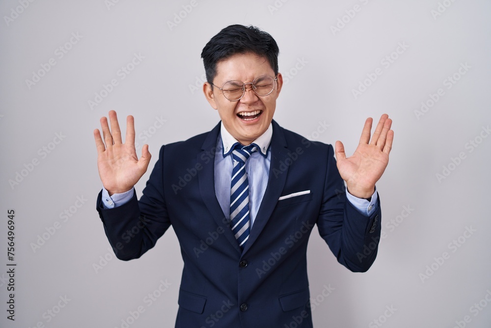 Young asian man wearing business suit and tie celebrating mad and crazy for success with arms raised and closed eyes screaming excited. winner concept Young asian man wearing business suit and tie celebrating mad and crazy for success with arms raised and closed eyes screaming excited. winner concept