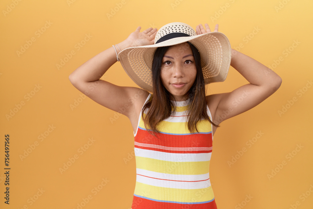 Middle age chinese woman wearing summer hat over yellow background doing bunny ears gesture with hands palms looking cynical and skeptical. easter rabbit concept. Middle age chinese woman wearing summer hat over yellow background doing bunny ears gesture with hands palms looking cynical and skeptical. easter rabbit concept.