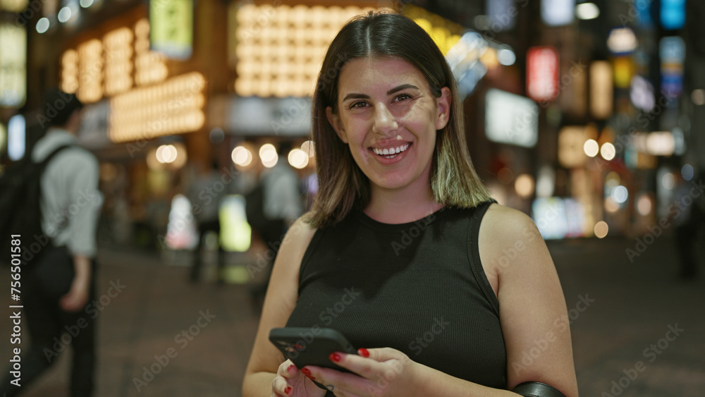Beautiful hispanic woman smiling at tokyo's urban night lights while texting happily on her smartphone Beautiful hispanic woman smiling at tokyo's urban night lights while texting happily on her smartphone