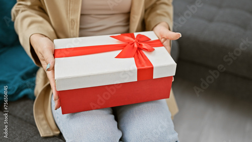 At home, a cozy comfort surrounds a woman sitting relaxed on a comfortable sofa, her hands gently holding a gift, adding charm to an otherwise ordinary living room