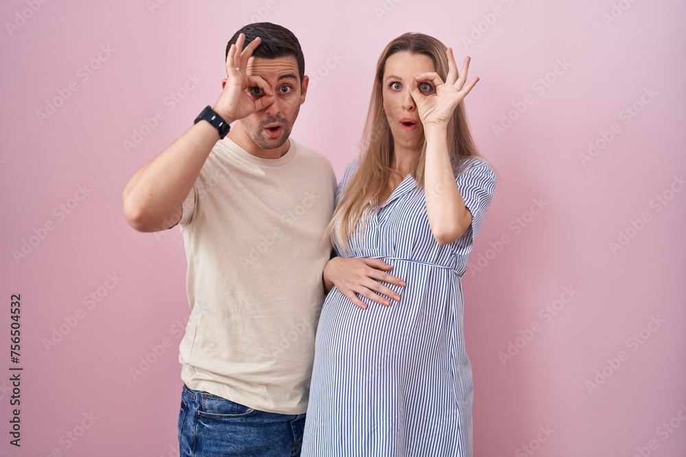Young couple expecting a baby standing over pink background doing ok gesture shocked with surprised face, eye looking through fingers. unbelieving expression. Young couple expecting a baby standing over pink background doing ok gesture shocked with surprised face, eye looking through fingers. unbelieving expression.