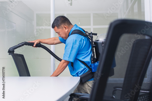 A professional janitor in blue uniform using a commercial backpack vacuum to clean a modern office chair in a bright workspace.