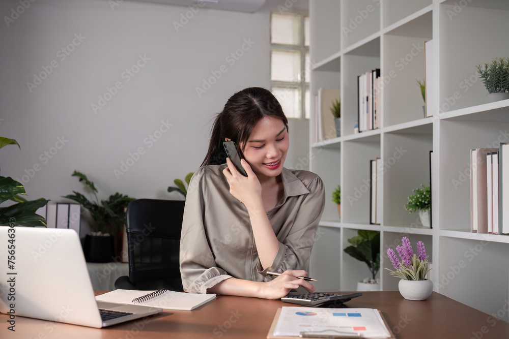 Young Asian business woman sits on the phone in an online business meeting using a laptop in a modern home office decorated with shady green plants. Young Asian business woman sits on the phone in an online business meeting using a laptop in a modern home office decorated with shady green plants.