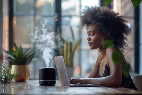 black essential oil diffuser on the table in the office, African girl is sitting with a laptop. Concept aromatherapy and relaxing. Air freshener photo