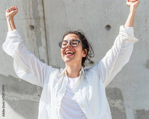 Victorious young woman with glasses raising her arms in jubilation against a concrete backdrop, capturing the hopecore essence of personal victory and pure joy photo