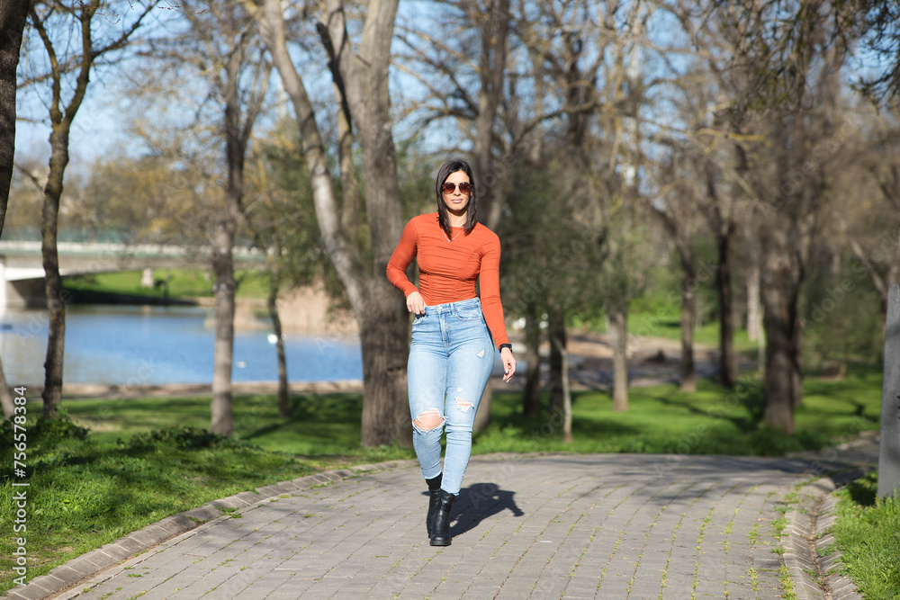 Fototapeta premium Young and beautiful brunette woman dressed in casual clothes and sunglasses, walks along a path between deciduous trees in autumn, in the background a lake.