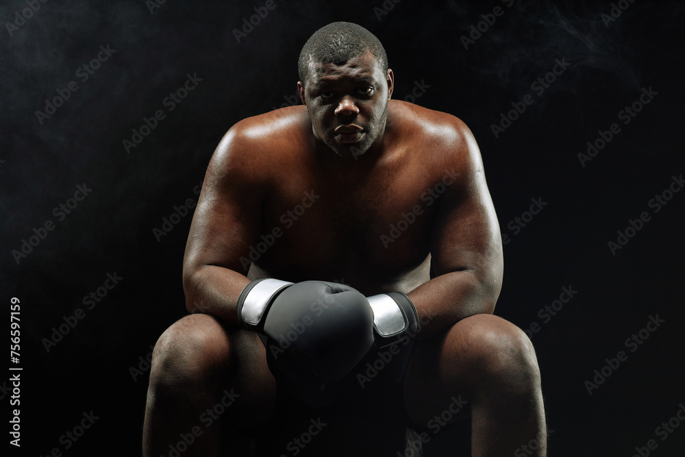 Dramatic backlit portrait of muscular African American boxer looking at camera sitting against black background