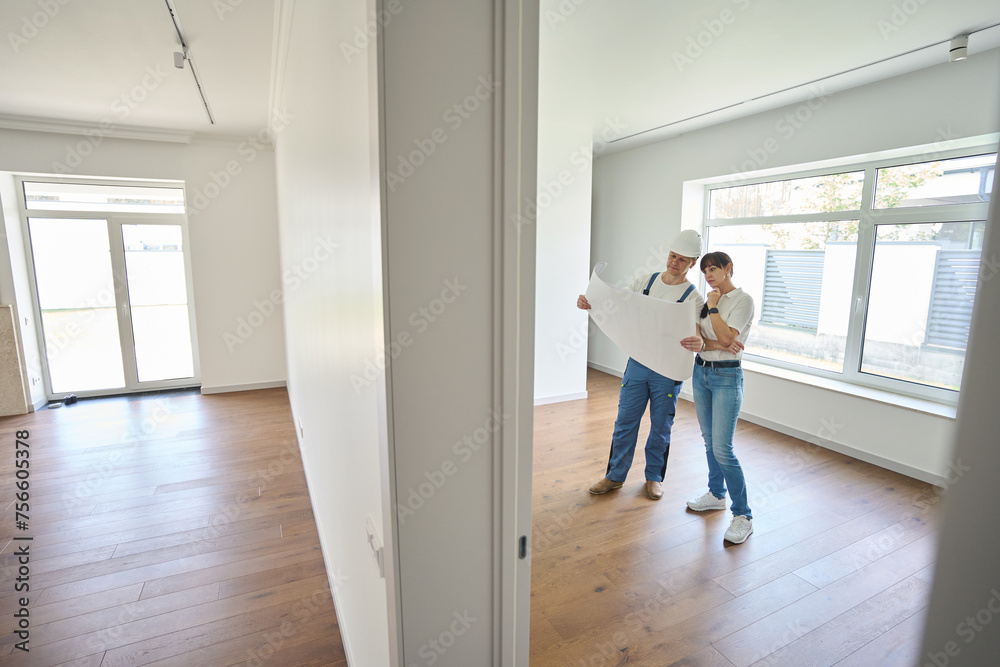 Focused male worker and woman watching blueprint in new townhouse before repair Focused male worker and woman watching blueprint in new townhouse before repair