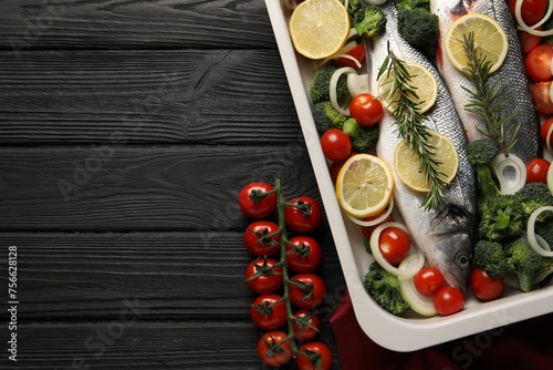 Raw fish with lemon in baking dish and vegetables on black wooden table, flat lay. Space for text