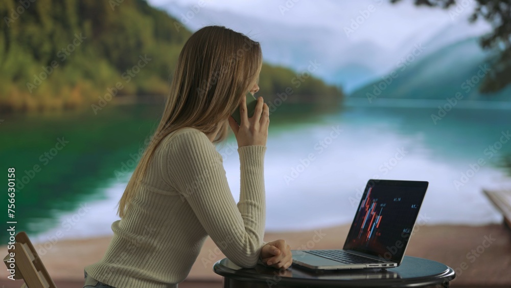 Female working from distance and travelling. Young woman freelancer working near mountain lake, talking on smartphone, looking at laptop.