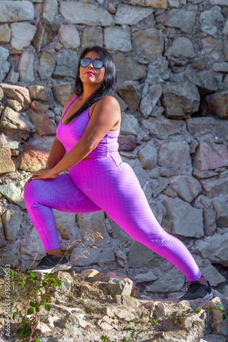 Sexy fat latin woman with positive and self-confident attitude, wearing tight pink sportswear, stretching her legs with her arms resting on her knees in the field. photo