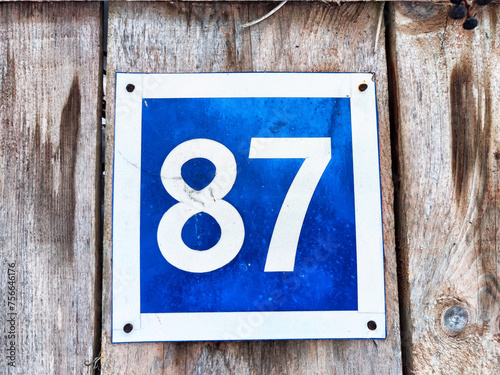 Number 87 on a Blue Sign Against Wooden Background. A bold white number 87 centered on a weathered blue square sign
