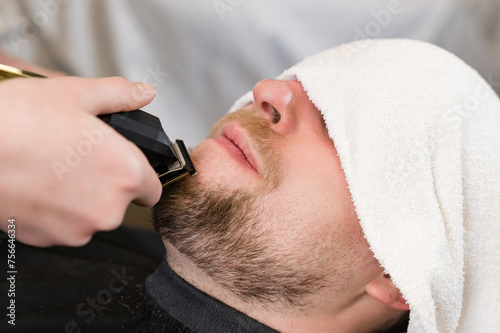 The clients head is covered with a towel during a beard trim. Shortening the length of the beard from the sides by the master for the client.