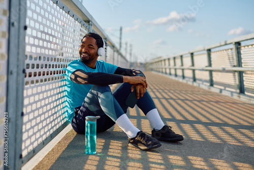 Happy black sportsman enjoying in music over headphones during water break outdoors.