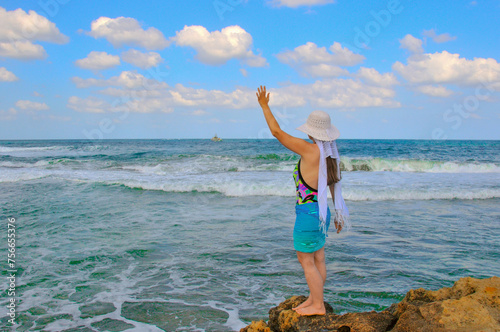 A girl standing on a rock waves her hand to a sailing ship in the sea