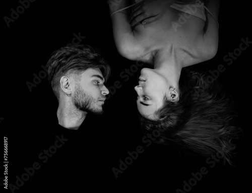 An overhead view of a man and woman lying together on a dark background and looking at each other. Black and white portrait.