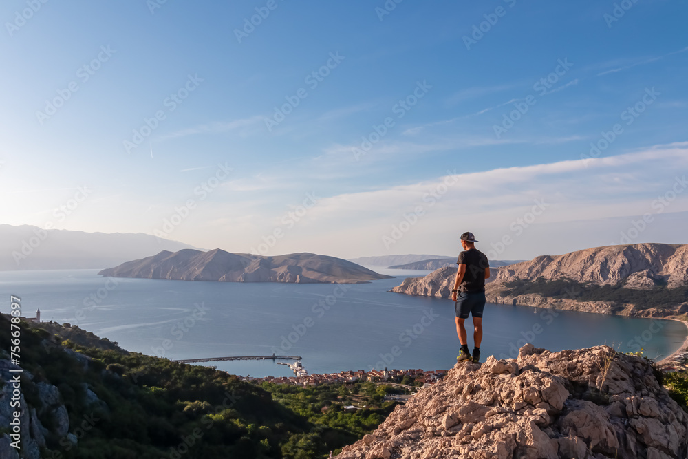 Fototapeta premium Hiker man on edge of rock cliff overlooking archipelago of Kvarner Bay in coastal town Baska, Krk Island, Primorje-Gorski Kotar, Croatia, Europe. Coastal hiking trail along coastline of Adriatic Sea