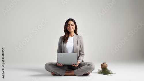 Woman sitting cross-legged on the floor, smiling and looking at a laptop placed on her lap against a plain white background.