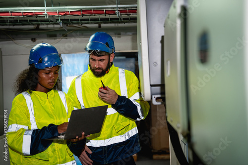 Two Engineers in yellow and blue safety gear are looking at a laptop. They are wearing hard hats and safety vests © NewSaetiew