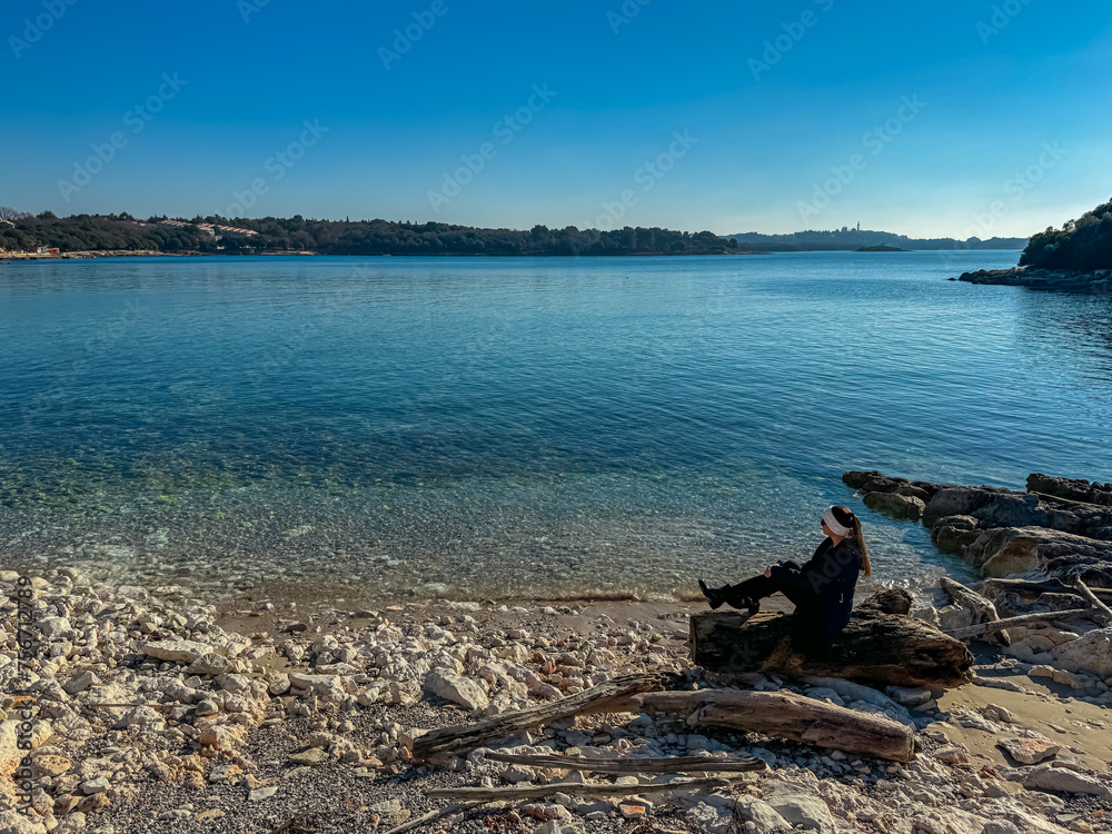 Woman sitting on idyllic pebble beach in coastal town Funtana, Istria, Croatia. Calm sea surface of Adriatic Mediterranean Sea in morning hours. Seaside vacation concept in summer. Tranquil atmosphere