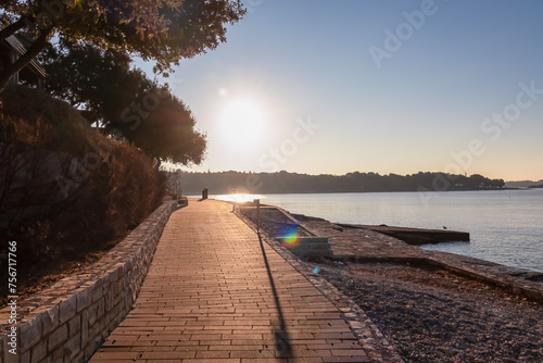 Fototapeta Naklejka Na Ścianę i Meble -  Idyllic promenade walkway along pebble beach of coastal town Funtana during sunset, Istria, Croatia. Calm sea surface and coastline of Adriatic Mediterranean Sea. Seaside vacation concept in summer