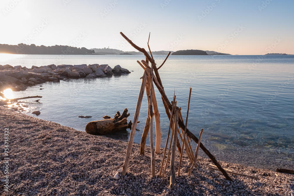Artwork made of wooden sticks on stone beach during sunrise in coastal village Funtana, Istria, Croatia. Distant view of Vrsar and tropical islands. Calm water surface at Adriatic Mediterranean Sea Artwork made of wooden sticks on stone beach during sunrise in coastal village Funtana, Istria, Croatia. Distant view of Vrsar and tropical islands. Calm water surface at Adriatic Mediterranean Sea
