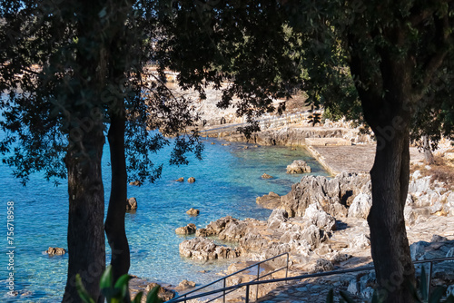 Idyllic pathway through forest with scenic view of green turquoise coloured bay in coastal town Funtana, Istria, Croatia. Rocky beach at Adriatic Mediterranean Sea. Seaside vacation concept in summer