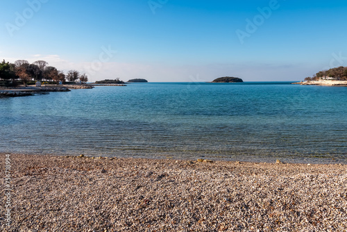Fototapeta Naklejka Na Ścianę i Meble -  Panoramic view of small islands seen from pebble beach in coastal town Funtana, Istria, Croatia. Calm sea surface of Adriatic Mediterranean Sea in morning hours. Seaside vacation concept in summer