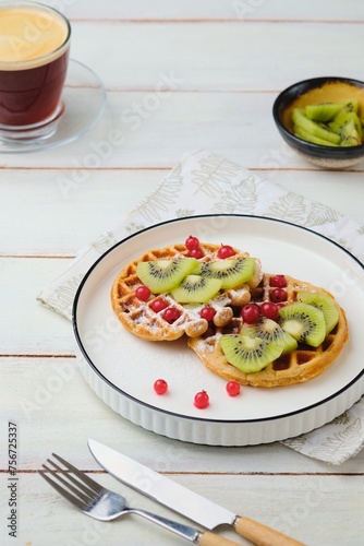 Two round waffles made of buckwheat flour, dusted with powdered sugar, topped with chopped fresh kiwi and red currants on a white plate on a light wooden background. Waffle recipes. Breakfasts.