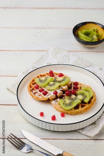 Two round waffles made of buckwheat flour, dusted with powdered sugar, topped with chopped fresh kiwi and red currants on a white plate on a light wooden background. Waffle recipes. Breakfasts.