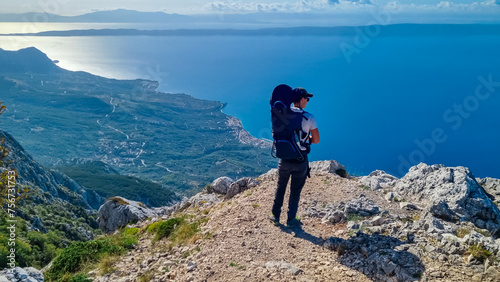 Father with baby carrier on top of mount Sinjal overlooking Biokovo nature park, Makarska, Dalmatia, Croatia. Hiking trail karst landscape. Dinaric Alps in summer. Coastline Adriatic Mediterranean Sea photo