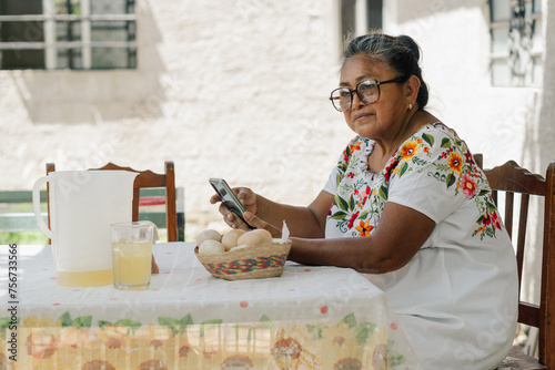 Elderly Latina woman sitting at home using a cell phone to send a text message.