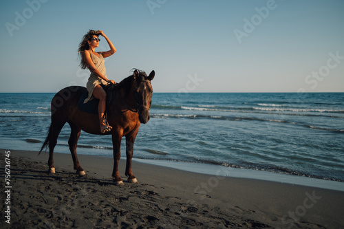 An attractive lady is posing on a horse near sea on a beach at sunset.